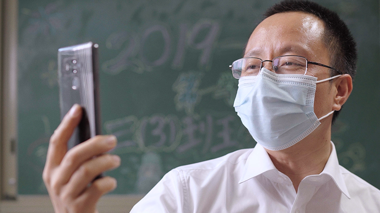 红英小学戴口罩班主任手持手机进行直播教学，黑板写着班级信息，疫情线上课堂